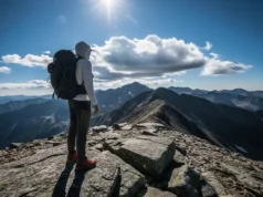 The Long Sleeve Shirt Layering Blueprint for Backpacking A full-body shot of a backpacker wearing a long-sleeve sun hoodie and a large pack, standing on a windy mountain ridge under a bright, partly cloudy sky.