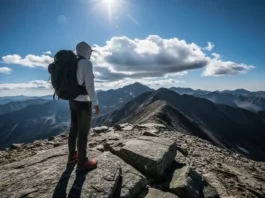A full-body shot of a backpacker wearing a long-sleeve sun hoodie and a large pack, standing on a windy mountain ridge under a bright, partly cloudy sky.