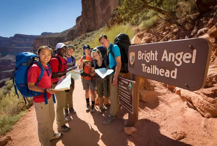 Hikers preparing at the Bright Angel Trailhead, Grand Canyon, with informational signs and the trail visible, illustrating a guide to parking and shuttles.