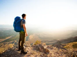 A full-body shot of a male hiker with a large backpack standing on a mountain peak, looking at the view on a sunny day.