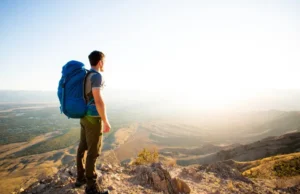 Men’s Hiking Bags Rated for Comfort: A 2025 Field Test A full-body shot of a male hiker with a large backpack standing on a mountain peak, looking at the view on a sunny day.
