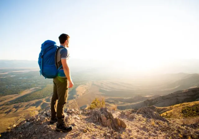 A full-body shot of a male hiker with a large backpack standing on a mountain peak, looking at the view on a sunny day.