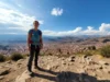 Women’s Walking Footwear: Packability & Terrain Tested A full-body shot of a woman in hiking gear standing on a mountain trail that overlooks a city in the distance, showcasing versatile footwear.