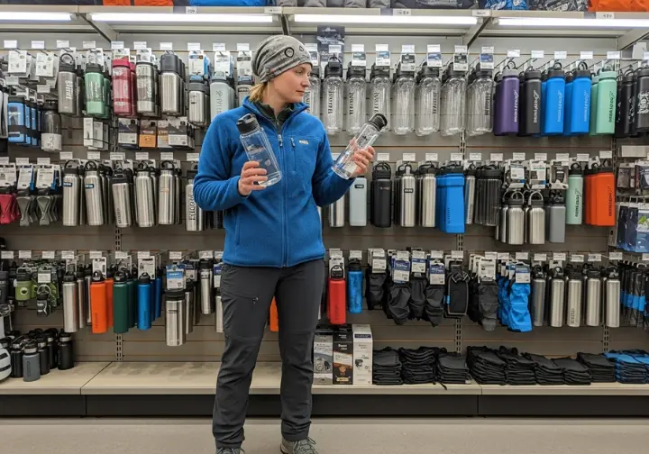 A hiker in a gear store looks at a wall of different water bottles, comparing a steel and a plastic one in their hands.