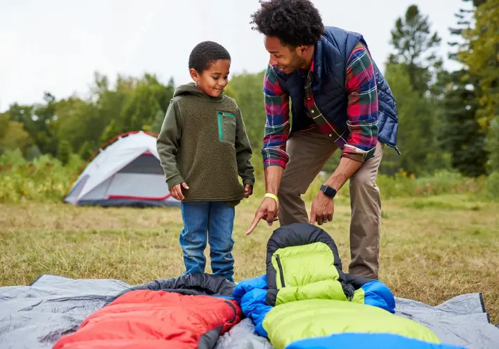A parent and child in hiking clothes stand over two children's sleeping bags laid out on a tarp at a campsite, with the parent explaining a feature.