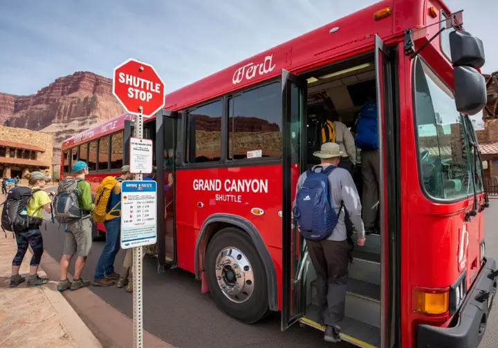 A red Grand Canyon shuttle bus at a busy stop with hikers, illustrating how to master the free shuttle system for accessing the Bright Angel Trailhead.