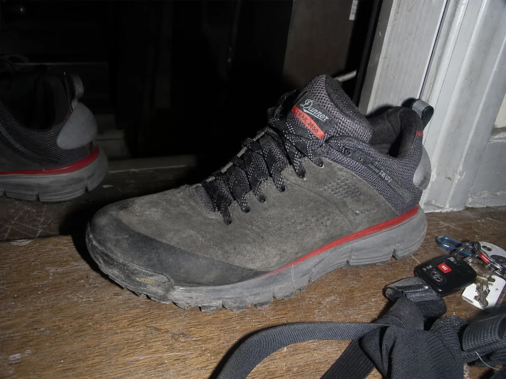 A worn suede low-cut hiking shoe by an apartment door showing months of trail-to-town scuffing