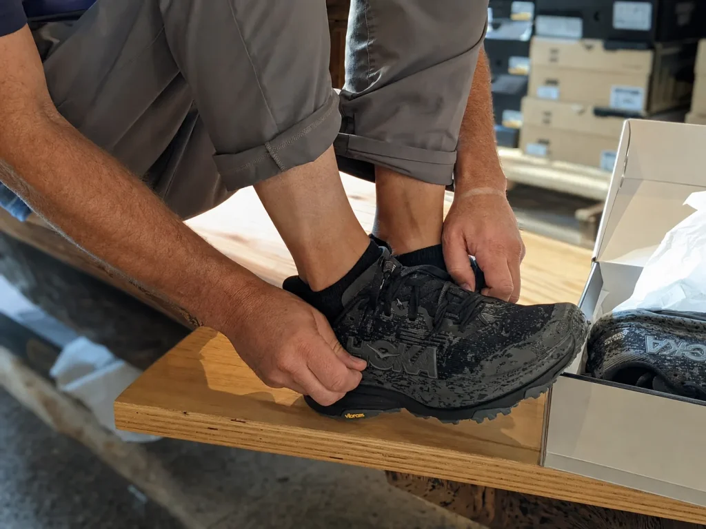 A hiker checking thumb-width toe fit while trying on a cushioned low-cut hiking shoe on a bench