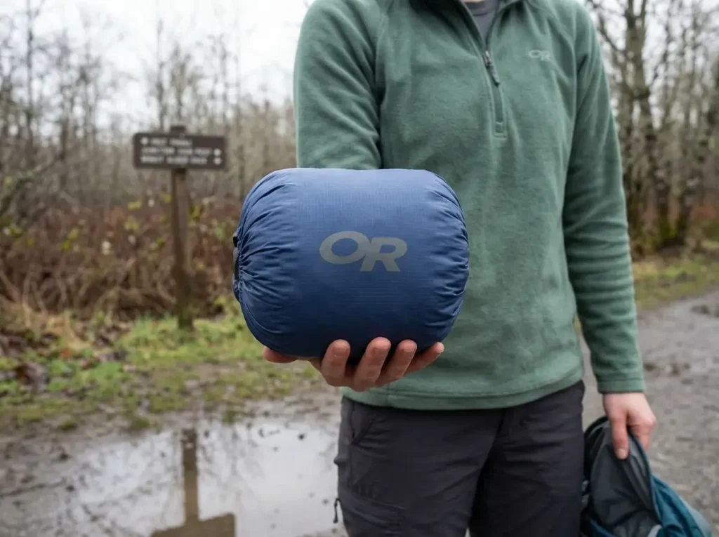 Packed Outdoor Research Helium rain jacket held in one hand beside an open daypack on the trail
