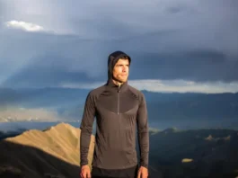 A hiker adjusts his long-sleeve hiking shirt layering system while standing on a mountain ridge with a view of a sunlit valley.