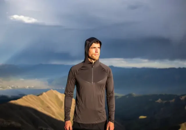 A hiker adjusts his long-sleeve hiking shirt layering system while standing on a mountain ridge with a view of a sunlit valley.
