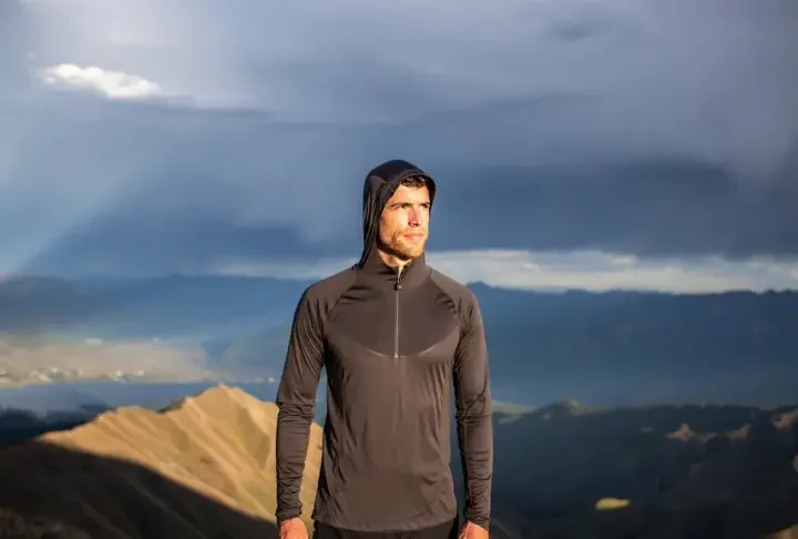 A hiker adjusts his long-sleeve hiking shirt layering system while standing on a mountain ridge with a view of a sunlit valley.