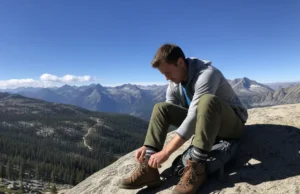 Trekking Socks Matched to Your Boots & Climate A full-body shot of a hiker sitting on a large rock on a mountain trail, tying their hiking boot, with a wool sock visible.