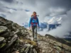 Best Women’s Hiking Coats: A Data-Backed Analysis A woman in a full-body shot wearing a technical hiking coat and backpack hikes on a rugged mountain trail under a dramatic, cloudy sky.
