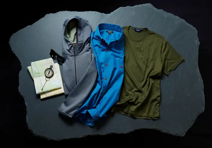 A flat-lay of the best men's hiking shirts of 2025, including a sun hoody, button-down, and merino tee, arranged on a slate surface with a map and compass.