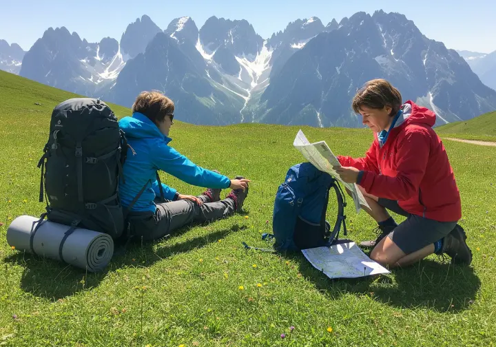 A full-body shot of two hikers with different styles of backpacks taking a break in a high alpine meadow with mountains in the background.