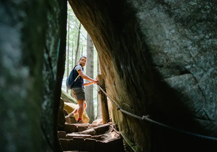 A male hiker passes through Arch Rock on the Alum Cave Trail using cable handrails, illustrating a mile-by-mile guide to the journey to the bluffs.
