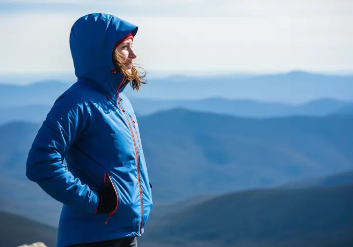 Full-body photo of a woman in a high-performance hiking jacket standing on a mountain peak, enjoying the panoramic view of valleys below.