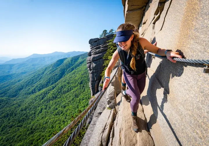 A female hiker traverses a narrow ledge with a cable handrail on the Alum Cave Trail, symbolizing the epic ascent to Mount LeConte beyond the bluffs.