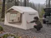 Best Inflatable Tents for Camping and Where They Fall Short Woman checking the air beam of a beige inflatable tent at a wooded car campsite on an overcast morning