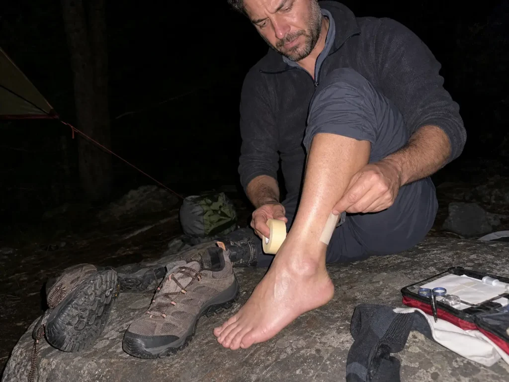 Hiker taping a hot spot on his heel at dusk beside his trekking shoes