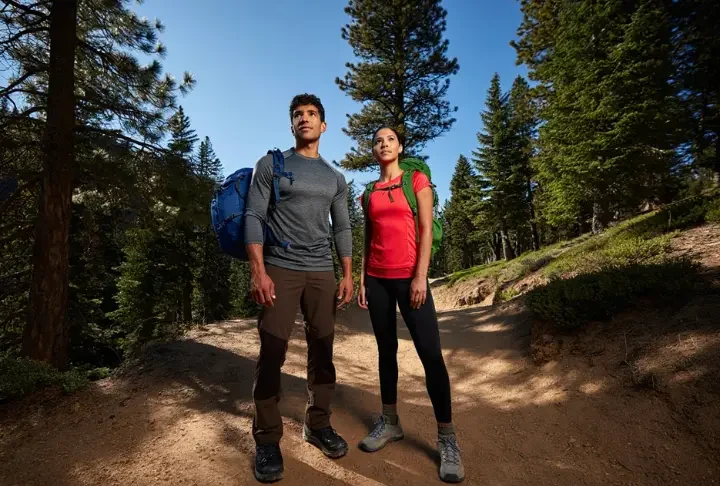 Two diverse hikers stand on a sunlit mountain trail, fully equipped with essential beginner hiking gear, embodying preparedness and confidence.