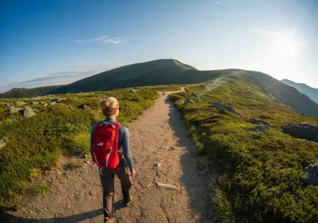 A hiker stands on a mountain path, gazing at a distant summit, representing the journey from first steps to achieving a hiking goal.