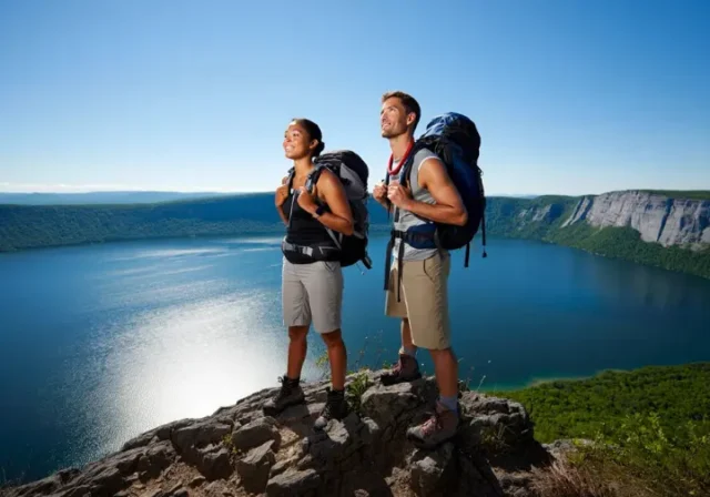 Two hikers stand on a rocky outcrop, gazing at the panoramic view of Minnewaska State Park Preserve, showcasing its diverse trails from lake loops to epic vistas.