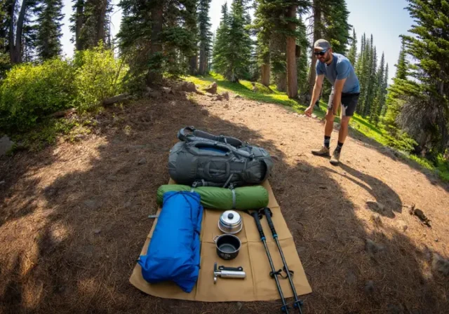A thru-hiker stands beside their organized gear spread on the ground, showcasing the "Big Three" and other essential items for a comprehensive thru-hiking gear list.