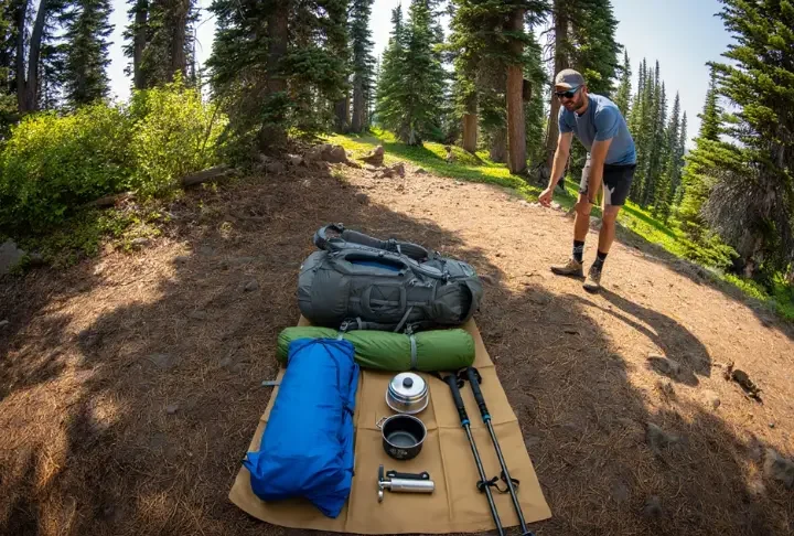 A thru-hiker stands beside their organized gear spread on the ground, showcasing the "Big Three" and other essential items for a comprehensive thru-hiking gear list.