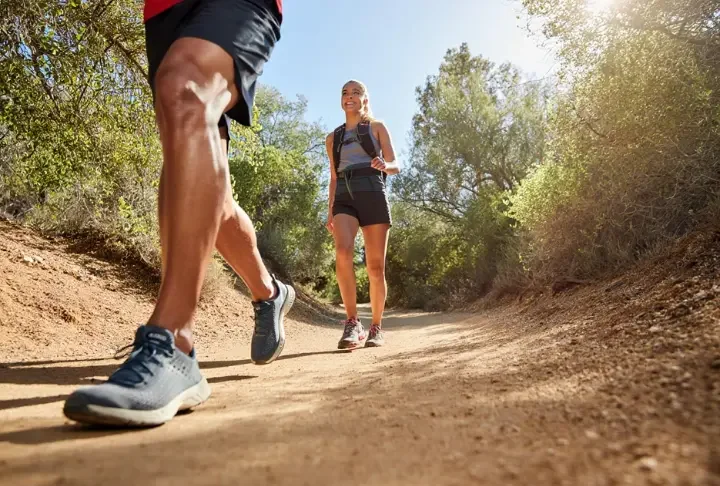 Two hikers on a trail, one in trail runners and the other in hiking shoes, visually representing the choice between different hiking footwear.
