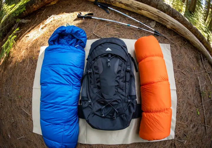 The "Big Three" thru-hiking gear items – backpack, tent, and sleeping bag – laid out in a forest, representing the core shelter and carry system for a thru-hiking gear list.