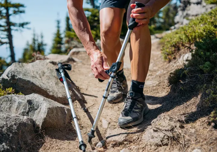 A hiker adjusts the length of their hiking pole on a steep, rocky trail for dynamic sizing based on varied terrain.