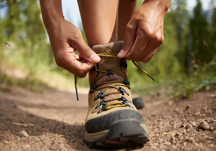Hands demonstrating a lacing technique on a hiking boot on a trail, illustrating how to solve common hiking pains and adjust fit.