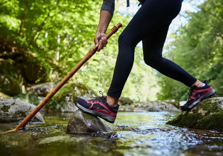 A hiker uses a handcrafted wooden staff for stability while crossing a rocky stream on a forest trail, demonstrating its practical use.
