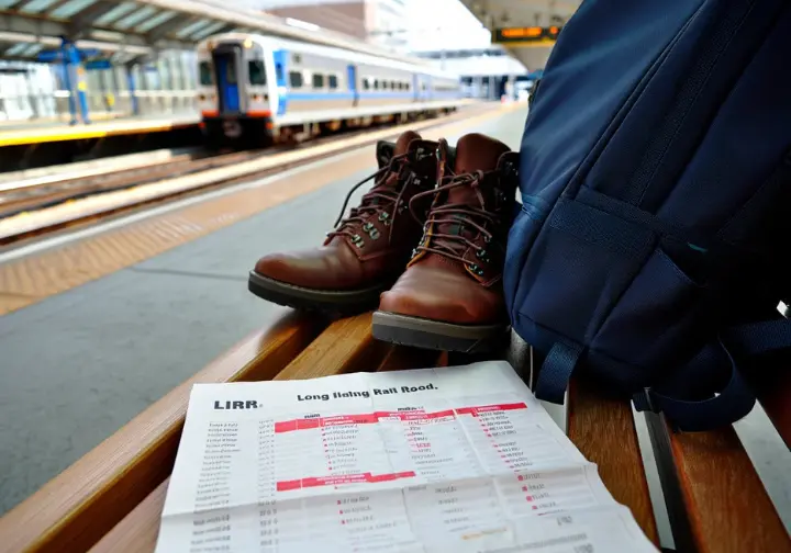 Hiking boots and a backpack next to a train schedule, with a train in the background, illustrating car-free hiking to places to hike on Long Island via public transit.