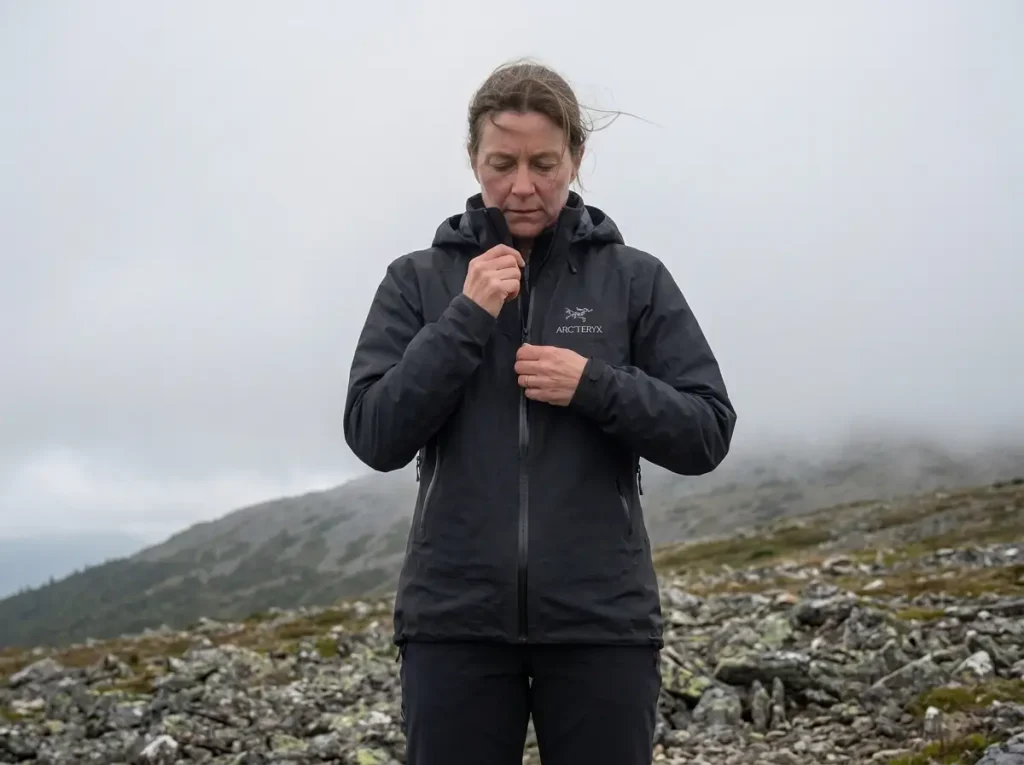 Hiker zipping a rain shell closed against the wind on an open above-treeline slope