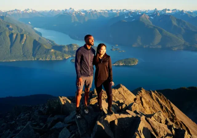 Two hikers watching the sunset from a mountain summit overlooking the epic coastal mountain scenery of Vancouver, BC.