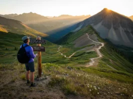 A hiker stands at a trail junction overlooking a panoramic vista of the Colorado Rockies at sunrise, with multiple trails of varying difficulty visible in the valley below.