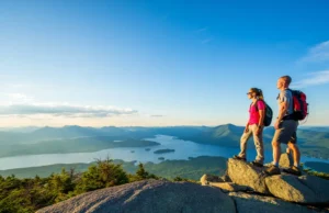 Conquer the Adirondacks: Lake George Hiking Guide Two hikers at a summit overlook, enjoying the panoramic view of Lake George and the Adirondack Mountains at sunrise.