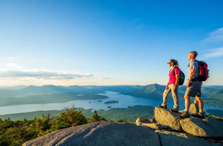 Two hikers at a summit overlook, enjoying the panoramic view of Lake George and the Adirondack Mountains at sunrise.