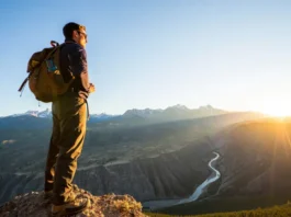 A hiker stands on a mountain overlook during golden hour, gazing at a vast American wilderness of canyons and peaks, representing the variety of epic trails in the US.