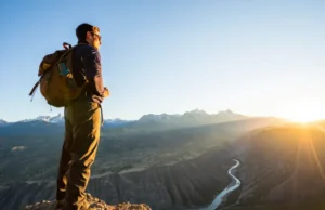 The Hiker’s Guide to 20 Epic US Trails (All Levels) A hiker stands on a mountain overlook during golden hour, gazing at a vast American wilderness of canyons and peaks, representing the variety of epic trails in the US.