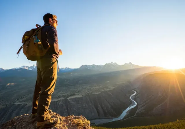 A hiker stands on a mountain overlook during golden hour, gazing at a vast American wilderness of canyons and peaks, representing the variety of epic trails in the US.