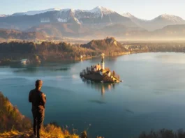 A hiker at the Ojstrica viewpoint looking at the iconic panoramic view of Lake Bled, Slovenia at sunrise, with the island and castle visible.