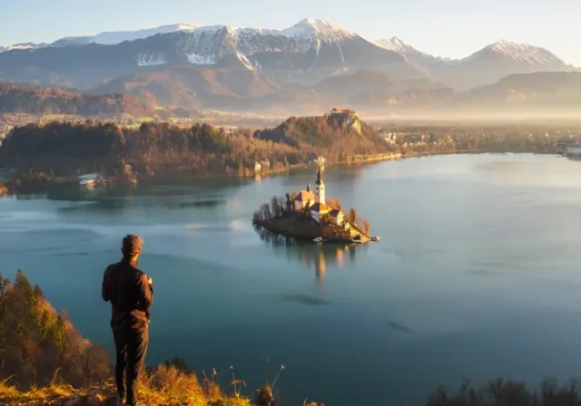 A hiker at the Ojstrica viewpoint looking at the iconic panoramic view of Lake Bled, Slovenia at sunrise, with the island and castle visible.
