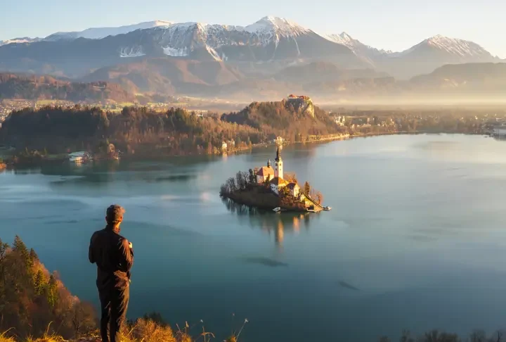 A hiker at the Ojstrica viewpoint looking at the iconic panoramic view of Lake Bled, Slovenia at sunrise, with the island and castle visible.