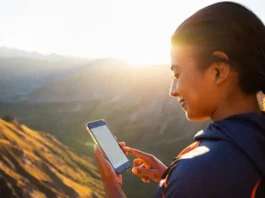 A hiker stands confidently on a mountain summit at golden hour, looking out at a stunning valley while checking a trail map on her smartphone.