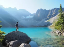A hiker stands before the stunning turquoise water of Upper Joffre Lake, looking towards the snow-covered mountains and glacier, a reward for getting a Joffre Lake day pass.