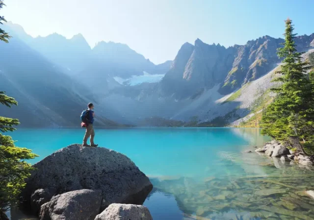 A hiker stands before the stunning turquoise water of Upper Joffre Lake, looking towards the snow-covered mountains and glacier, a reward for getting a Joffre Lake day pass.